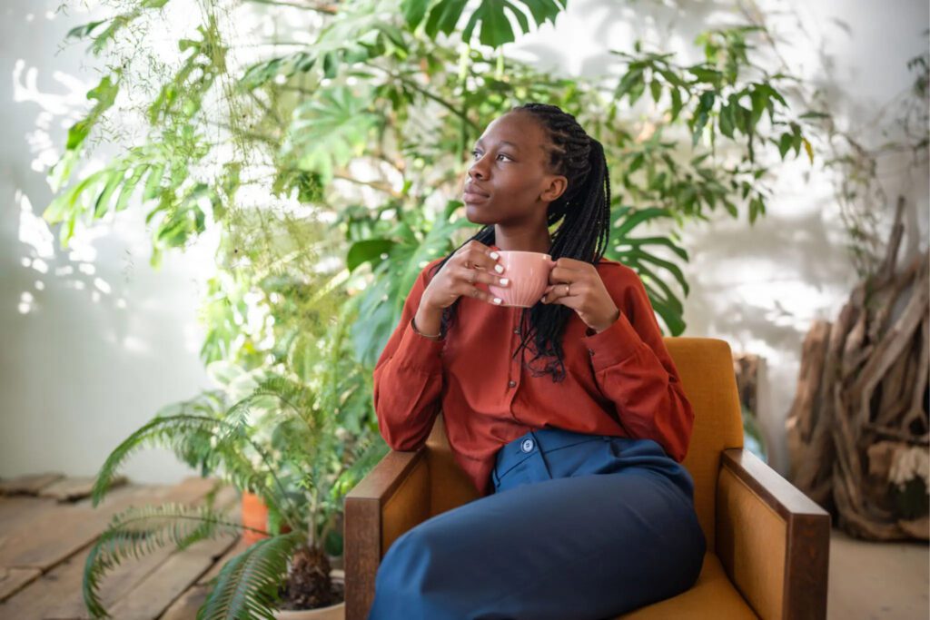 A woman in a rust-colored blouse sits on a mustard chair, holding a cup near a leafy plant.